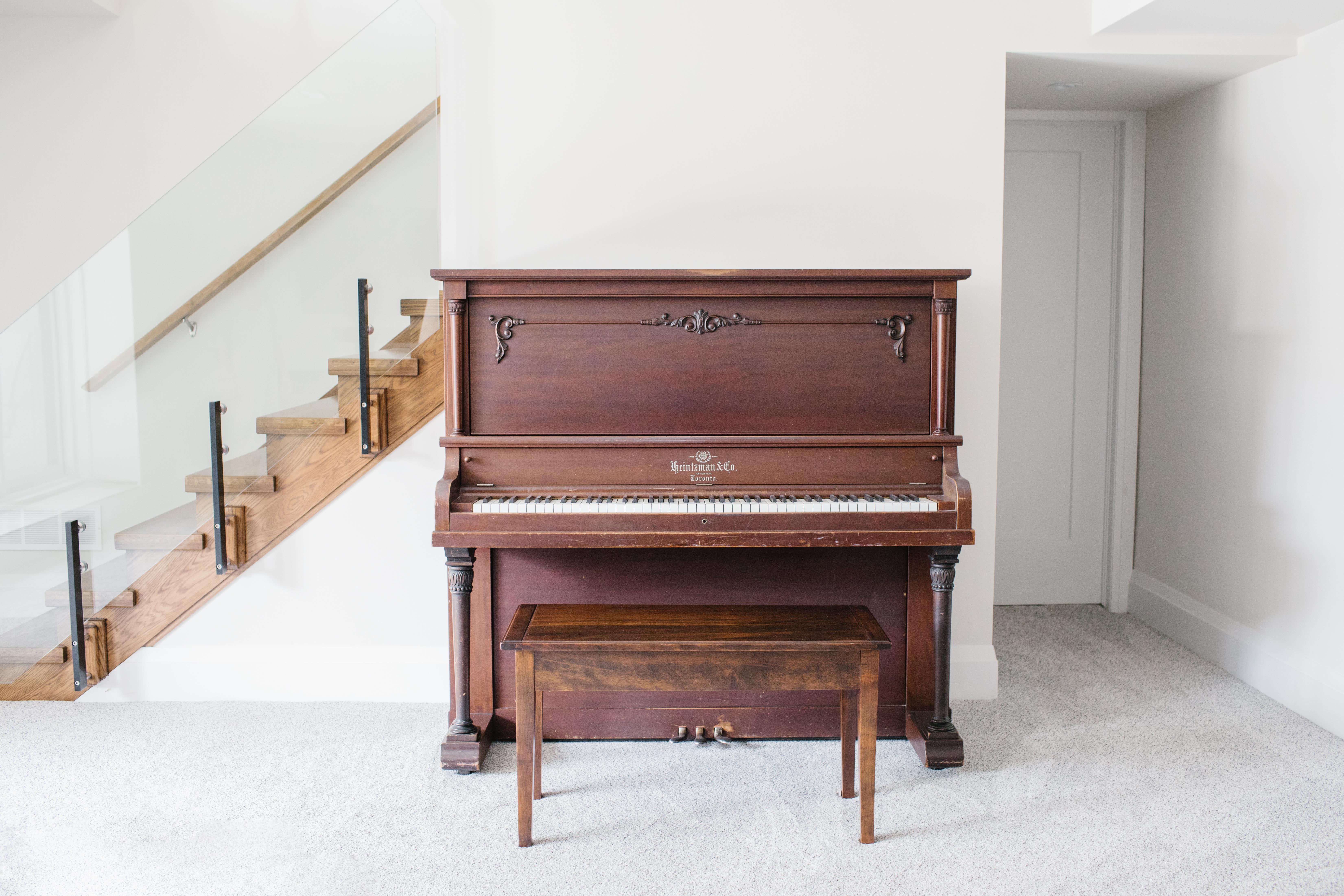 A beautiful antique Heintzman piano sits against a cream coloured wall at the base of a glass staircase. The piano lines up meticulously with the wall.